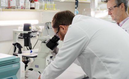 a man bends to look into a microscope at a container of yellow liquid watched on by another man, both are in white coats